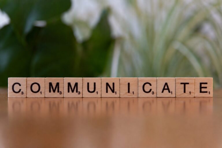 Fa Scrabble-kockákból kirakott COMMUNICATE szó zöld növény előtt, a hatékony kommunikáció témáját szimbolizálva